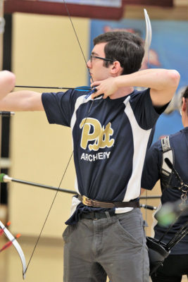 Young man in a Pitt Archery Club jersey at full draw with a recurve bow