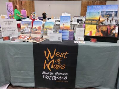 Picture of a table with a grey tablecloth and a black table runner that says "West of Mars Susan Helene Gottfried" with a display of Susan's books on top